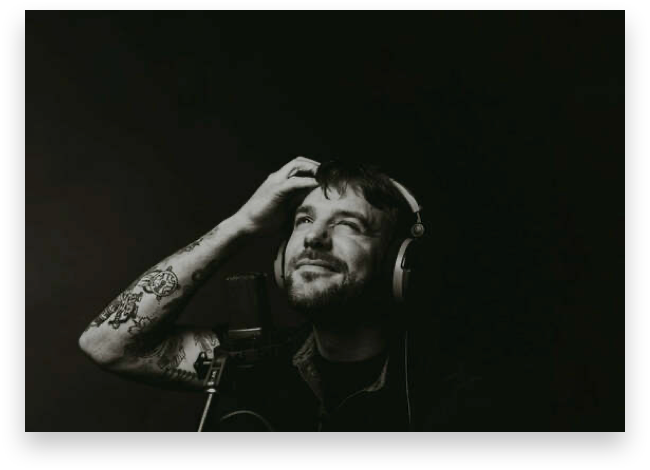 A guy with a beard and shaggy hair wearing headphones a looking a bit perplexed while standing in front of a mic