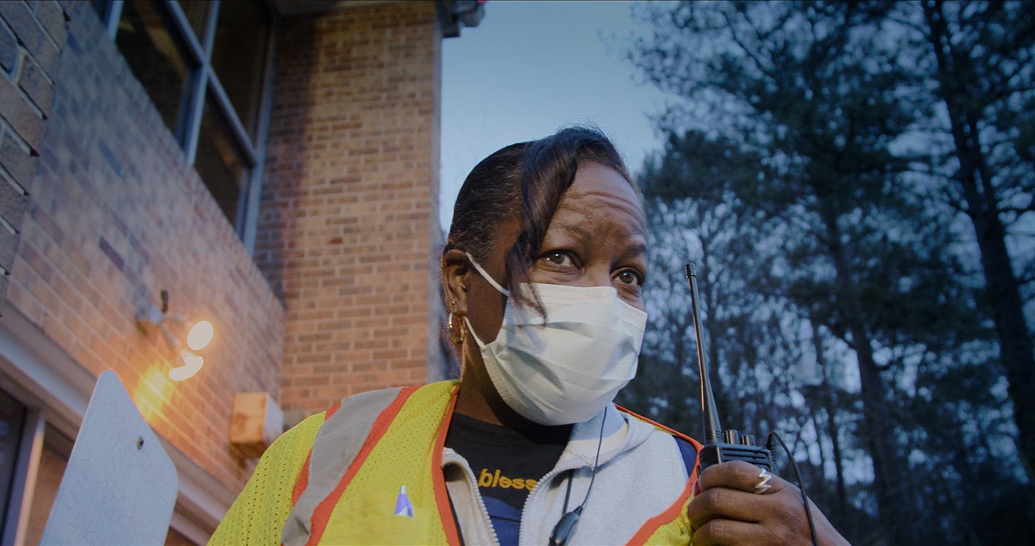 A Black woman wearing a surgical mask and a neon yellow vest talks into a walkie-talkie, while standing outside of a brick building.