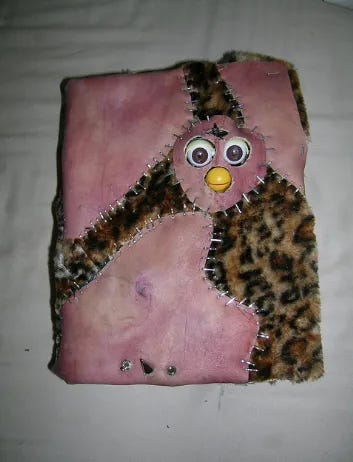 A pior imagem que deus abençoe você não vai ver na sua vida. Foto dessas tiradas com flash à noite no lençol branco tentando fazer ele parecer um fundo neutro. É a capa de um caderno fechado com uns pedaços de furby, o boneco amaldiçoado dos anos 90, costurado. Esse é foda de explicar, mas vamos lá. É uma energia Frankenstein. Tem uns três pedaços de algo que se assemelha a um couro muito desgastado num rosa claro já fodido. Daí tem meio que um pedação em um tecido peludo de oncinha. Todos os tecidos estão presos com uma porrada de grampos, tipo, grampeador, e tem os óio e o bico mecânico do furby saindo pra fora do caderno. Tem também um pino meio punk que pode ou não ser uma alusão a um pênis no inferior da página. tudo é horrível, extremamente amaldiçoado.