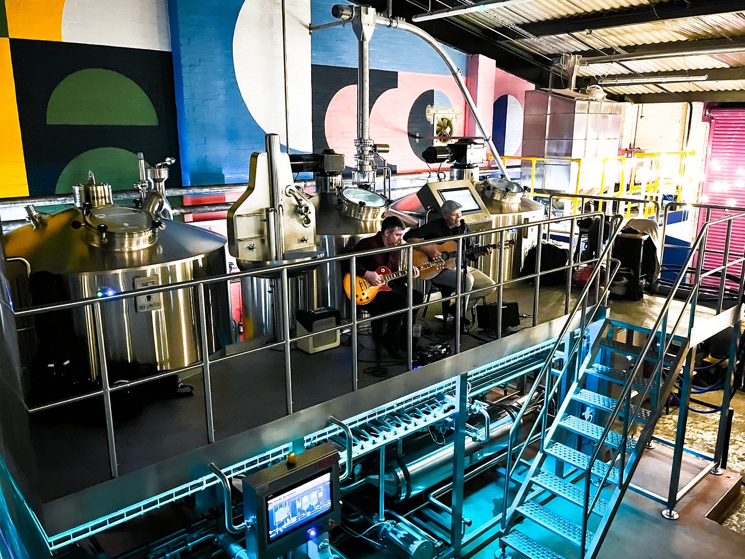 Two men perform with guitars in front of the brewery equipment inside.