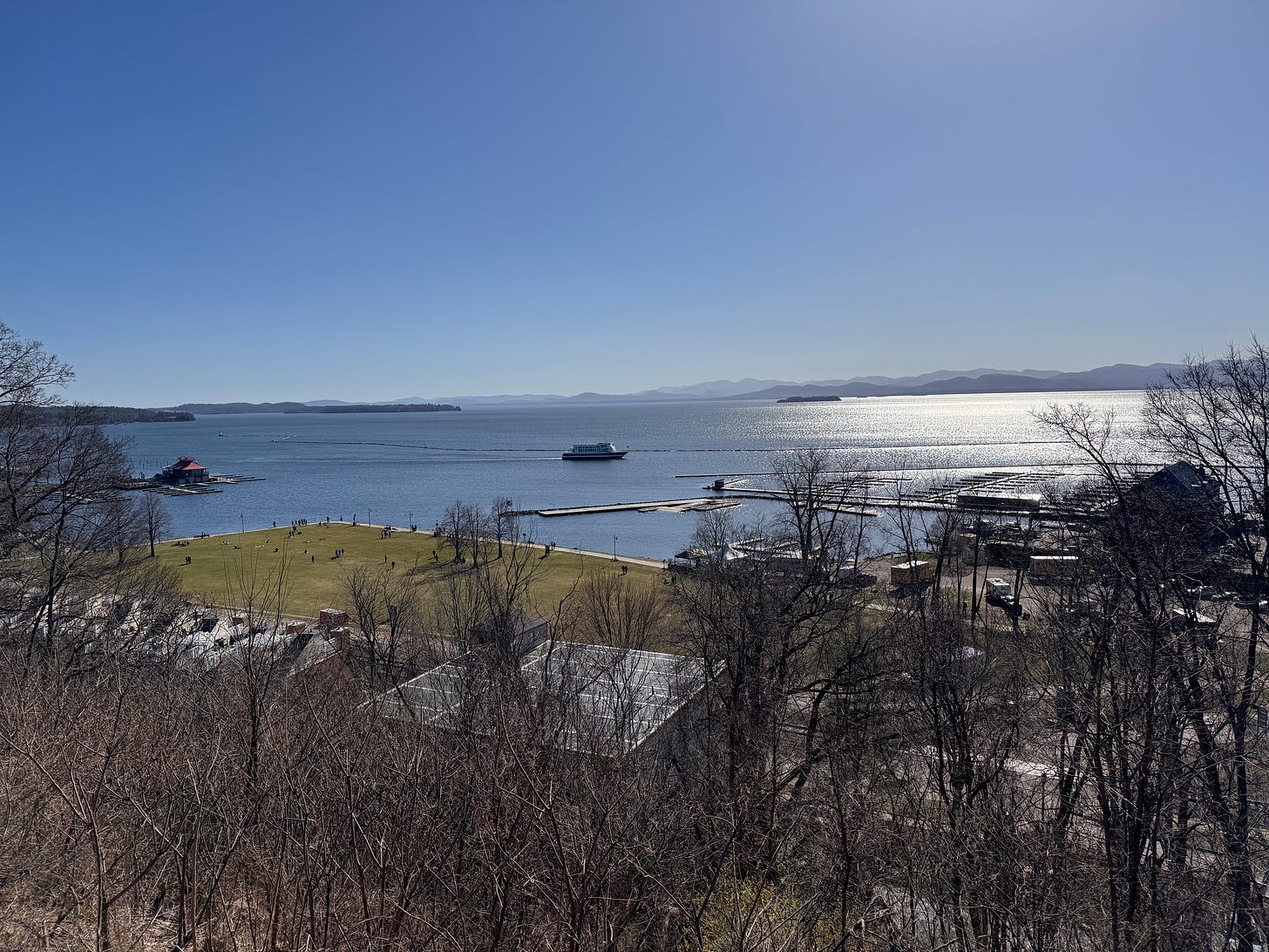 Lake Champlain stretches out from the shore of Burlington, a large boat in the foreground. The Adirondack mountain range is in the background, and the waters shimmer on a perfectly clear day.