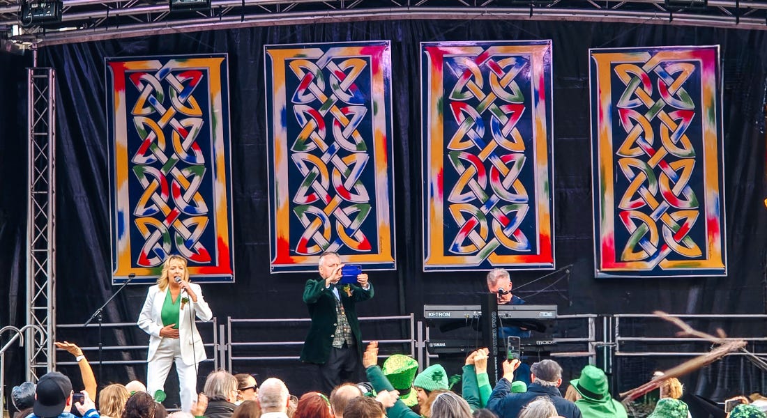 A stage with a black backdrop, three people on stage, and a crowd of people in green outfits below them A stage with a black backdrop, three people on stage, and a crowd of people in green outfits below them