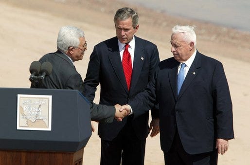 George Bush looking on as two men shake hands in a desert. no one seems to be having fun