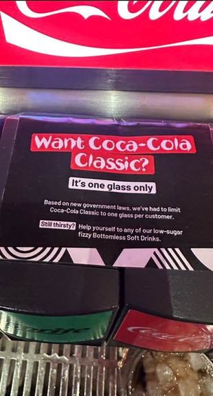 Coca-Cola dispenser machine in a store setting with red and black branding, featuring a sign on the front panel that reads Want Coca-Cola Classic? Its one glass only based on new government laws to help sell other low-sugar drinks youd be limiting, next to green low-sugar drink options and a drain below.