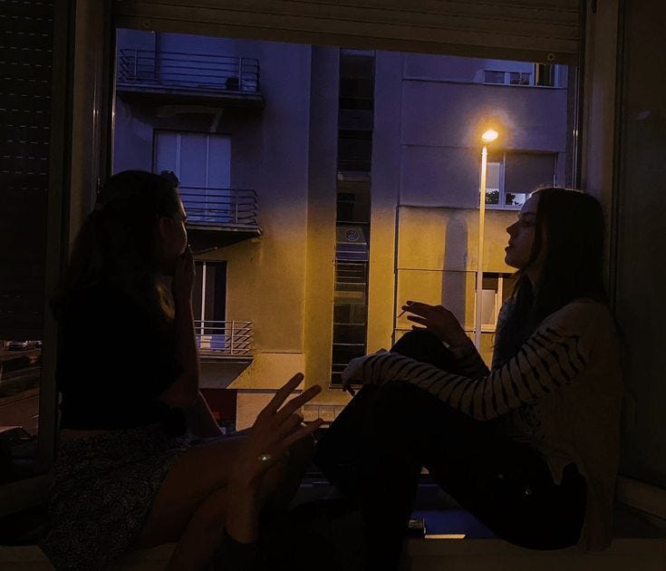 This may contain: two women sitting on a window sill talking to each other in front of a street light