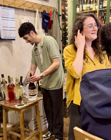 Photos inside In Vino wine bar in Yerevan, including a hand holding a bottle of Trinity Canyon Areni Noir, staff pouring wine behind shelves stocked with Armenian and international bottles, friends toasting with glasses of white wine, a long tasting table set with glassware beneath a sign reading 'The Renaissance of Armenian Wine Making,' and a wall of photos honoring winemakers