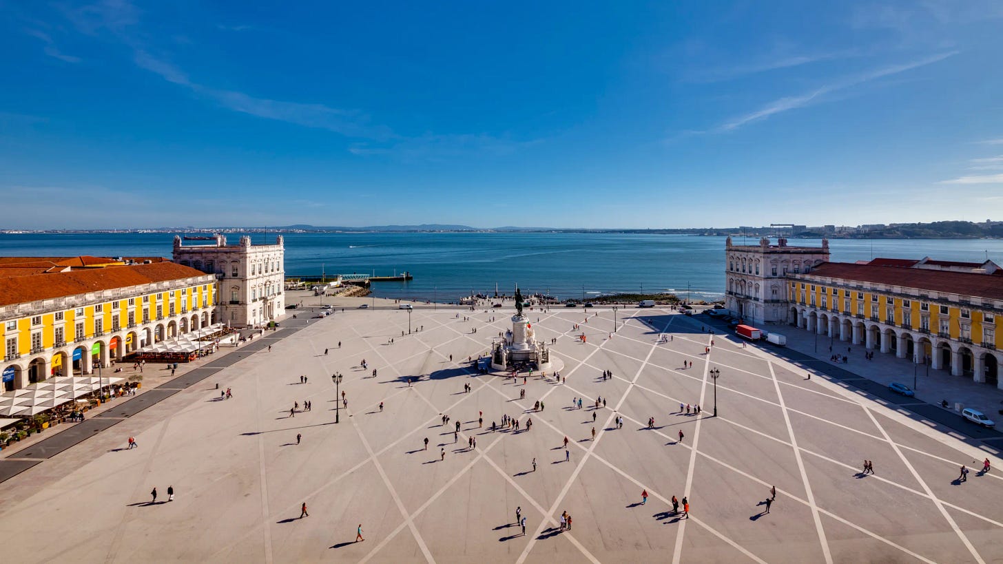  Una veduta aerea di Praca do Comercio a Lisbona, Portogallo, sotto un cielo azzurro e limpido. La piazza, di forma quadrata, è prevalentemente di colore chiaro, con un disegno di linee radiali nere che si estendono da un monumento centrale. Numerose persone sono sparse per la piazza, alcune sedute ai tavoli all'aperto lungo i bordi degli edifici che fiancheggiano i lati sinistro e destro. Questi edifici sono grandiosi e di colore giallo-arancio con tetti rossi e molte finestre. Al centro della piazza si erge una statua scura di una figura a cavallo. Oltre la piazza, in lontananza, si estende un vasto specchio d'acqua blu, con una costa collinare visibile all'orizzonte. Il cielo è di un blu brillante, senza nuvole.