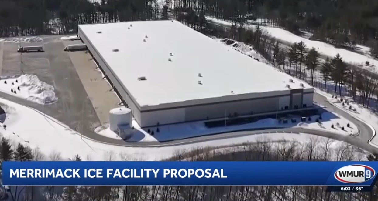 aerial shot of a large warehouse building in the snow; its parking lot and the road leading to it have been cleared of snow, but only two semi-trucks are parked in it, otherwise it appears vacant. 