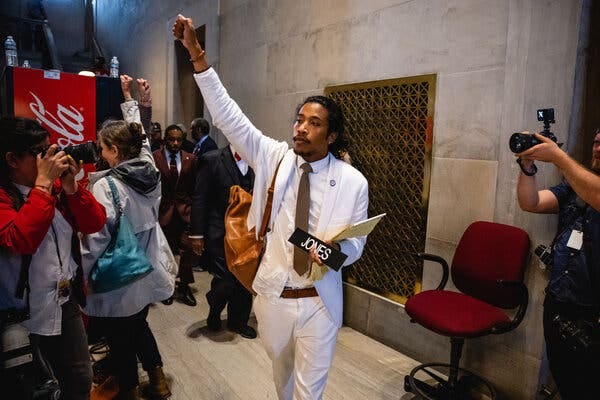 Representative Jones, in a white suit, white shirt and tan tie, leaves the House chamber, his arm hand raised high. He carries his desk nameplate. 