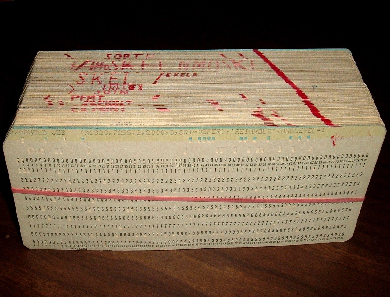 A stack of vintage punch cards photographed against a black background. The cards appear to be computer punch cards from an early computing era, with a light beige or manila color. The top card has handwritten red text that appears to include 'FORTRAN' and other programming-related notations. The cards show the characteristic rectangular holes punched in precise patterns across multiple rows, with numbered columns (0-9) repeated across the width of each card. The punch cards are stacked several inches thick, suggesting this represents a complete program or substantial dataset from the era of mainframe computing when programs and data were stored on these perforated cards.