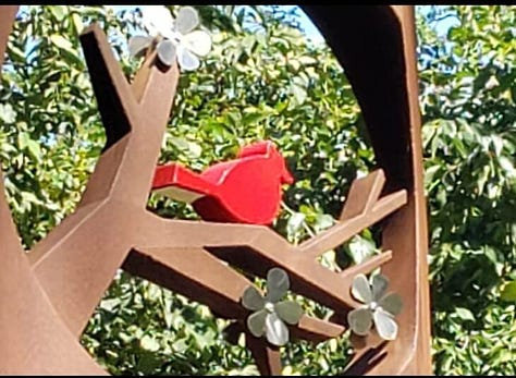 A cardinal mosaic in a shop with "please ask for assistance" under it, a cardinal in a tree sculpture in a park, and a cardinal stencil on a silver car door in a parking lot.