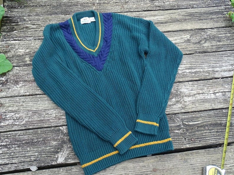 May include: Teal V-neck sweater with a cable knit design. The collar and cuffs have yellow and navy blue accents. The collar has a navy blue cable knit design with a yellow trim. The cuffs also have a yellow trim. The sweater is made of a thick, ribbed knit material.