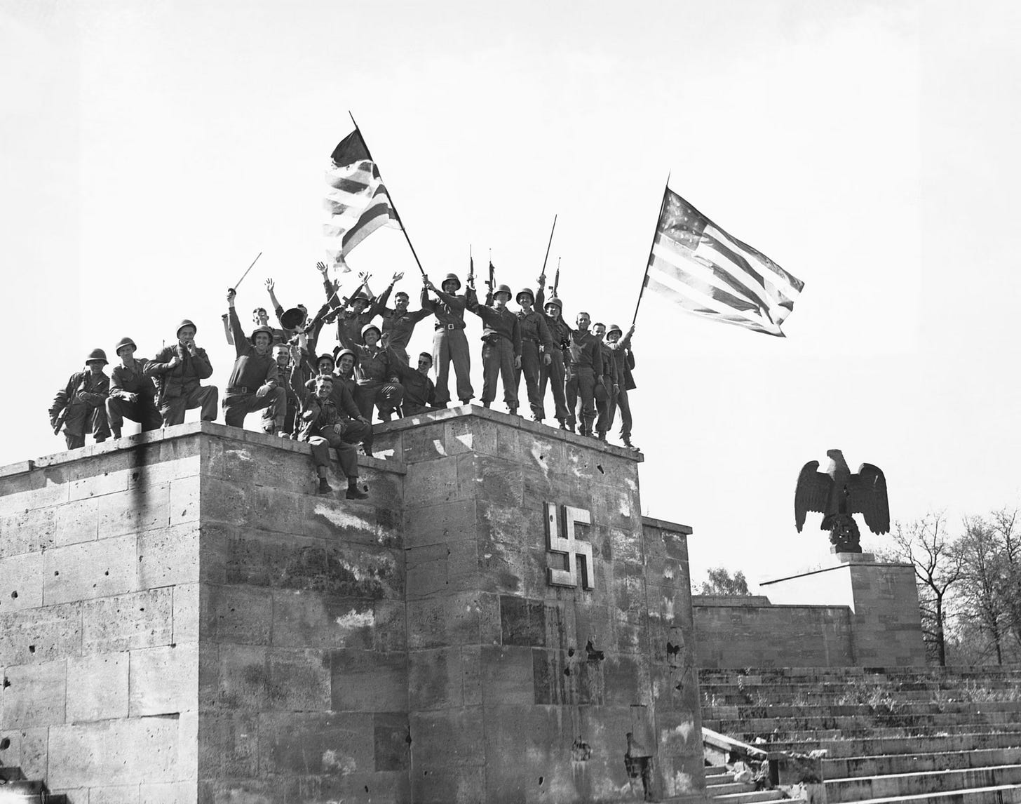 Historical photograph of WWII soldiers: This is a black-and-white historical photograph showing American soldiers celebrating on a stone structure adorned with a large Nazi swastika. The soldiers are raising American flags and their rifles in victory, likely at the end of World War II in Germany. This appears to be the Zeppelin Grandstand on the Nazi party rally grounds in Nuremberg.