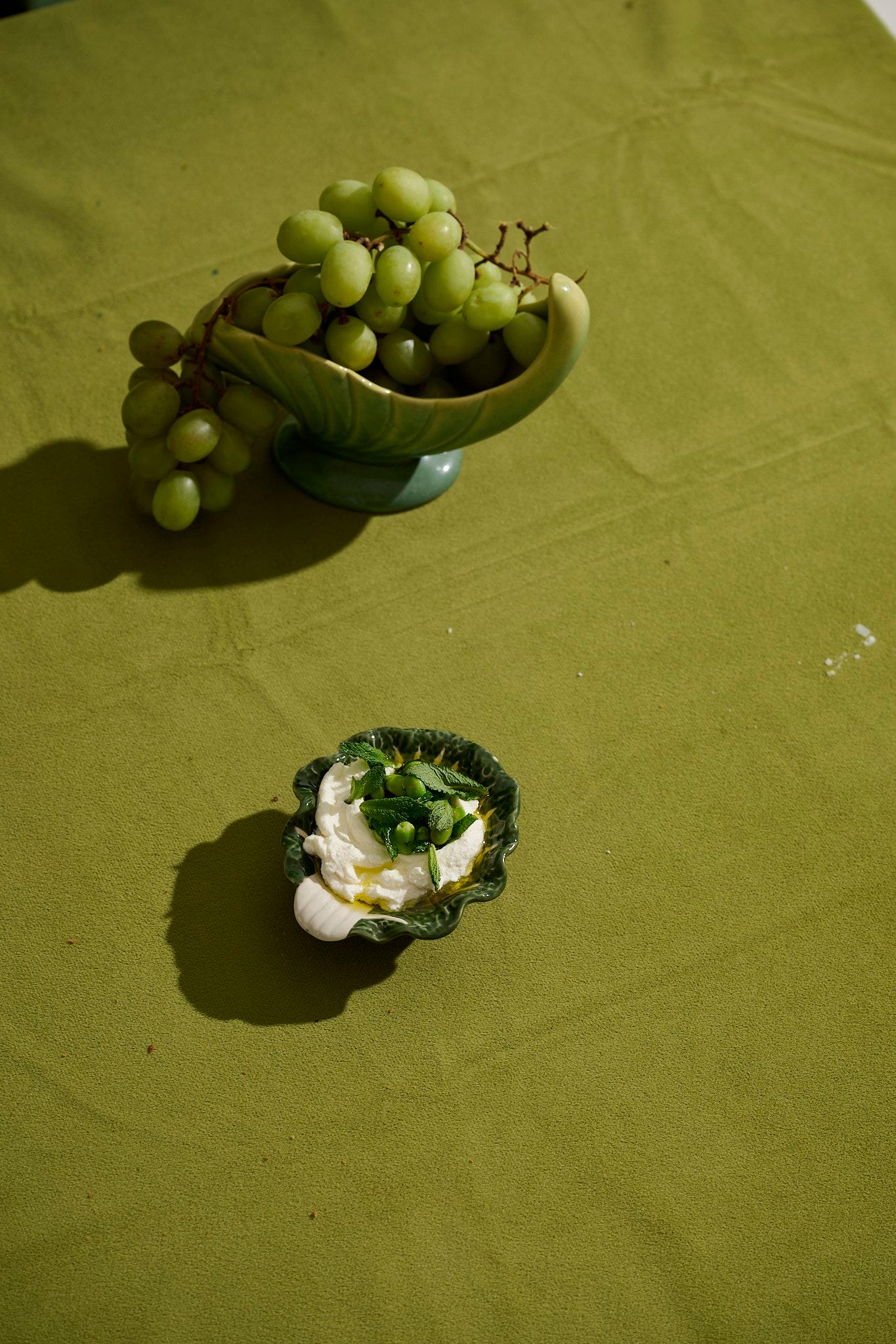 Whipped ricotta with fava beans, fresh mint, and olive oil served in a green cabbage-leaf dish, alongside green grapes in a vintage ceramic bowl on a green tablecloth.