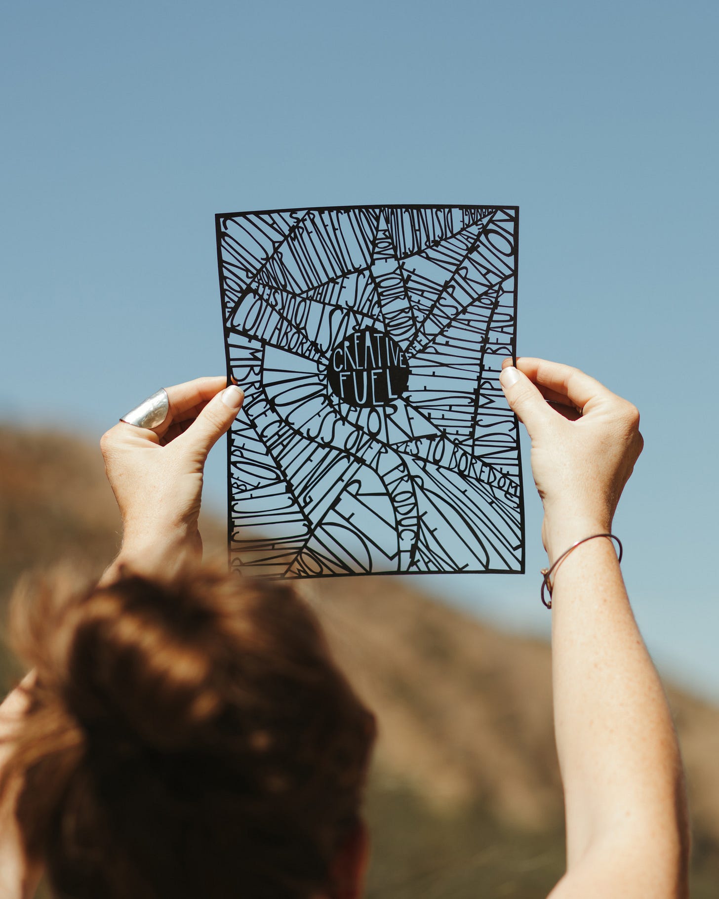 A photo of an elaborate paper cut piece of artwork being held up to a blue sky, with the words creative fuel carved out of the middle of it