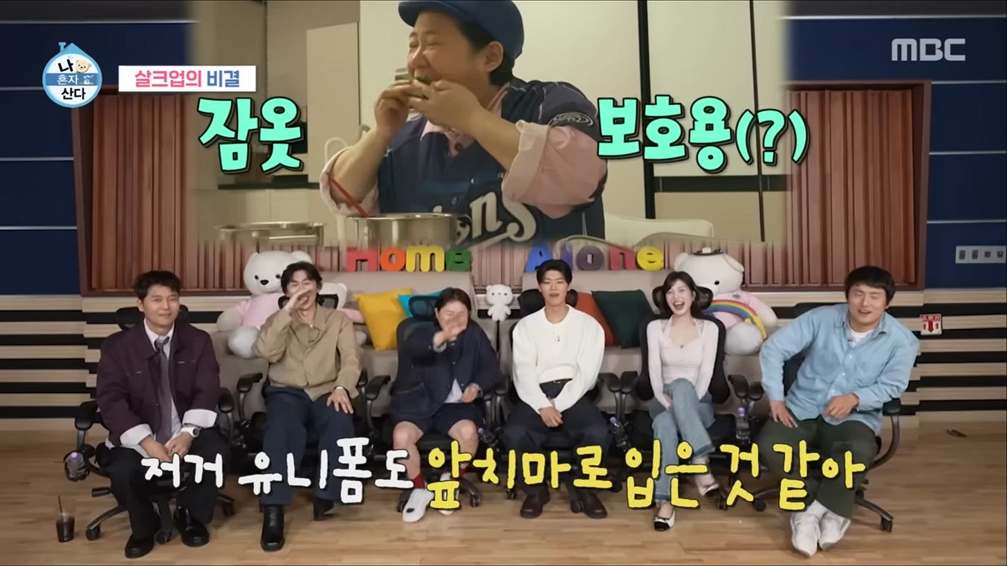 A television screenshot from the Korean reality show I Live Alone featuring a split-screen layout that illustrates the show's unique commentary format. On the right side of the screen, a celebrity is captured by a fixed camera inside his apartment, sitting at a simple table and eating a solo dinner. On the left side, the footage is framed within a studio setting where a panel of other celebrities sits together on a couch, pointing at the screen, laughing, and providing energetic commentary. The visual contrast perfectly captures how contemporary Korean television transforms private, solitary moments into a shared, public spectacle. A television screenshot from the Korean reality show I Live Alone featuring a split-screen layout that illustrates the show's unique commentary format. On the right side of the screen, a celebrity is captured by a fixed camera inside his apartment, sitting at a simple table and eating a solo dinner. On the left side, the footage is framed within a studio setting where a panel of other celebrities sits together on a couch, pointing at the screen, laughing, and providing energetic commentary. The visual contrast perfectly captures how contemporary Korean television transforms private, solitary moments into a shared, public spectacle.