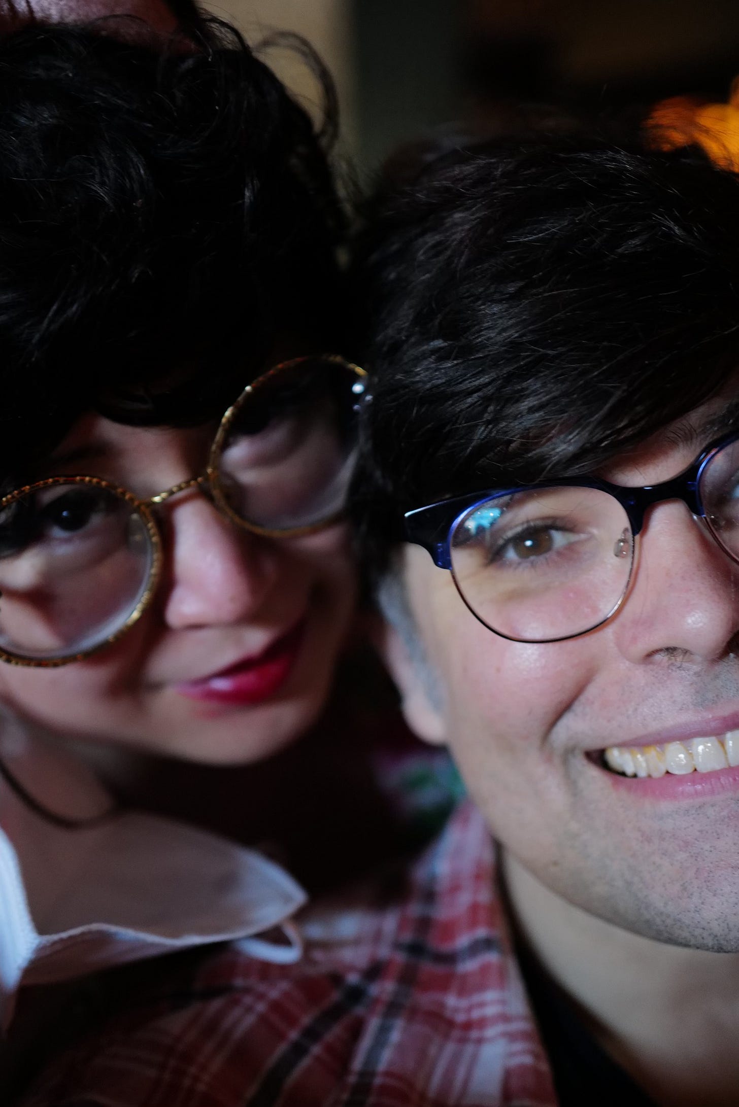 a woman with gold-rimmed glasses and black hair. She is an Asian woman smiling with her face pressed up against the face of a light-skinned man with glasses, who is also smiling. They are both exuberantly happy.
