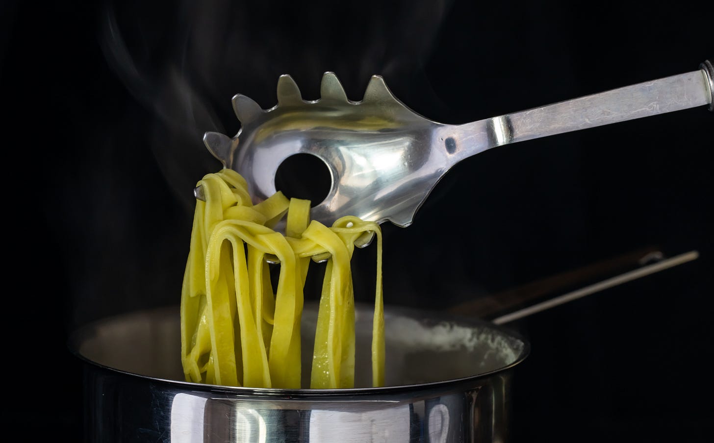 Metal pasta spoon with a central hole lifting flat noodles from a steaming pot against a dark background