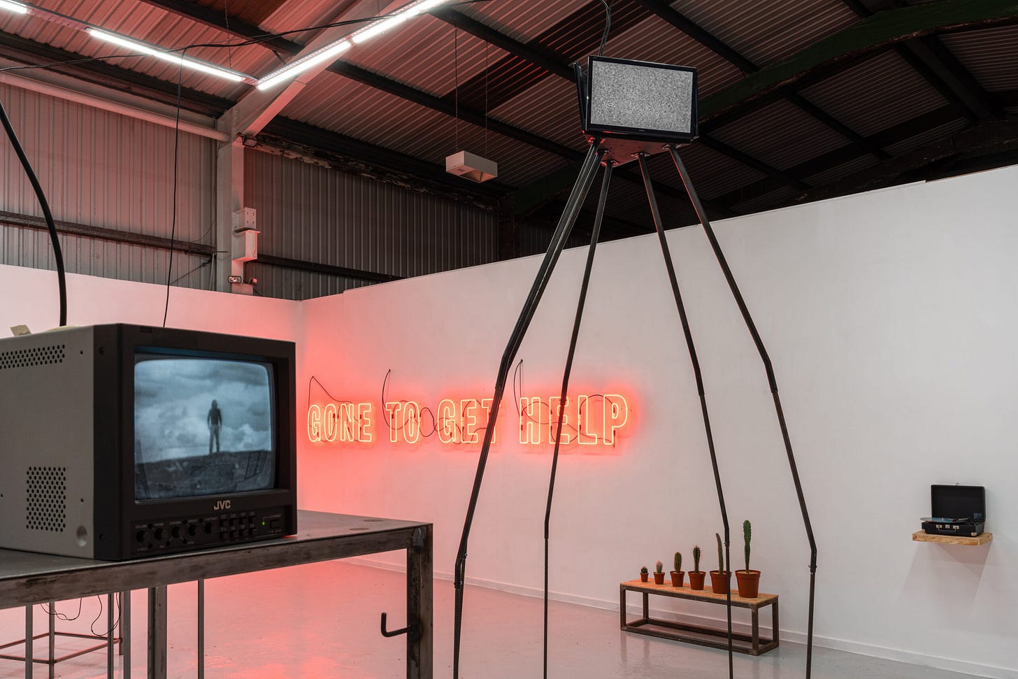 The image shows an installation view of the exhibition 'Two Plus Two Makes Four' at The Auxiliary project space in Middlesborough. A large wall-mounted work in bright orange neon strip lighting reads 'GONE TO GET HELP' and sits just above a bench supporting 6 cacti arranged in ascending order. In the foreground two small monitor screens sit atop welded steel mounts.