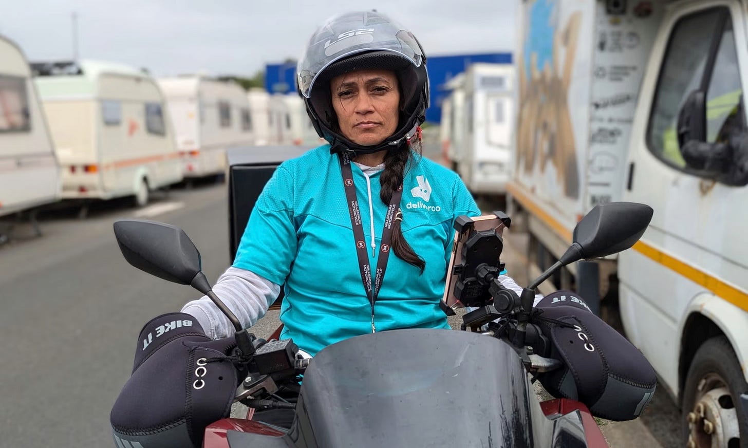 A fatigued woman wearing a bright turquoise Deliveroo top over their clothes, on their moped staring straight ahead at the viewer. A fatigued woman wearing a bright turquoise Deliveroo top over their clothes, on their moped staring straight ahead at the viewer.