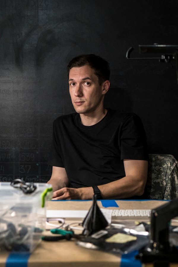 A young man sits at a cluttered desk and looks up with eyebrows slightly raised. A young man sits at a cluttered desk and looks up with eyebrows slightly raised.