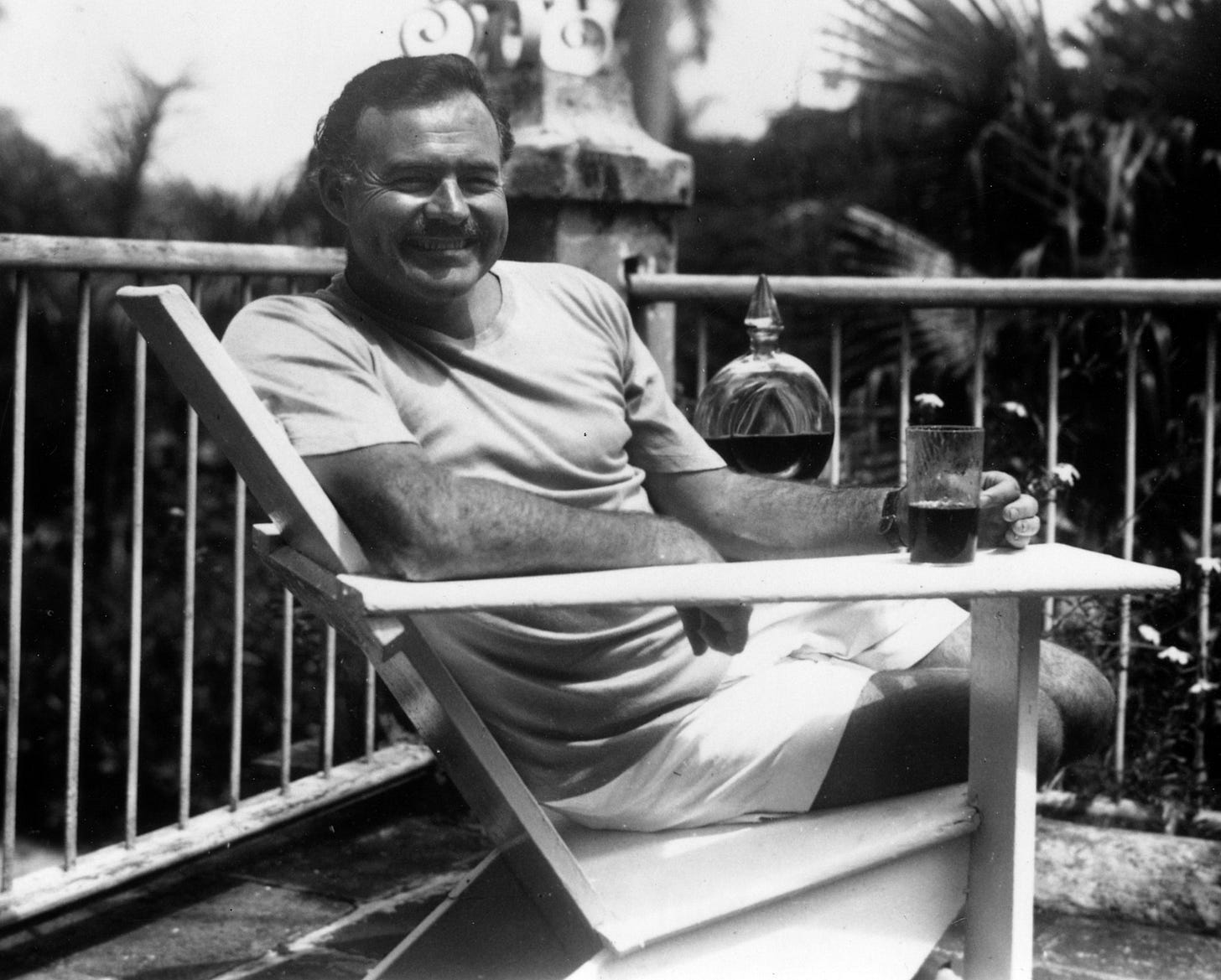 A man with short hair and a mustache is sitting in a chair outdoors, smiling, with a glass and a bottle on a table beside him. A man with short hair and a mustache is sitting in a chair outdoors, smiling, with a glass and a bottle on a table beside him.