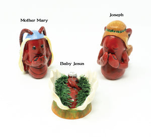 three lobster replicas labeled Mother Mary, Joseph, and Baby Jesus. Mary is wearing a blue headdress of some sort with her antenna sticking up through it and appears to be praying. Joseph is wearing a tan headpiece and has his eyes closed, also praying. Baby Jesus is lying down in a bed of what looks like green garnish.