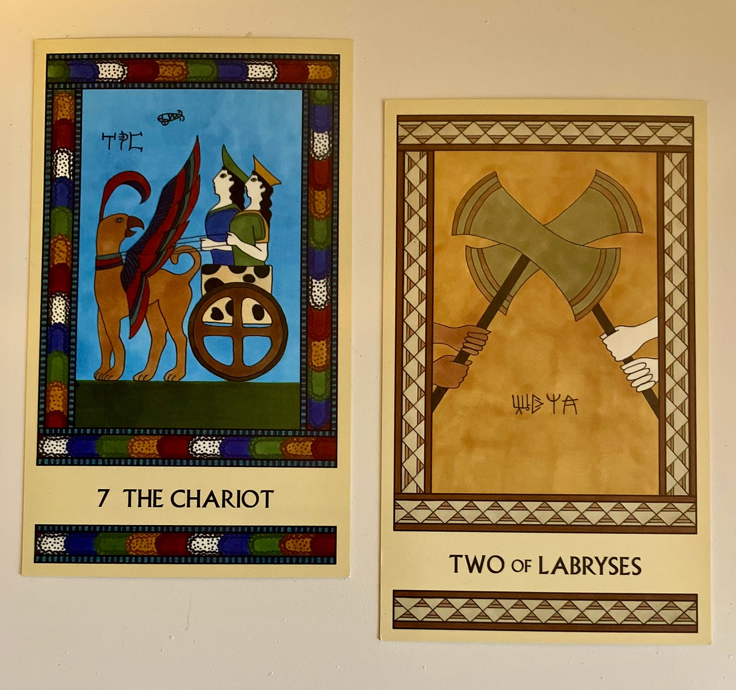 Two Minoan Tarot cards side by side on a cream surface. The Chariot, on the left, has a multicolored border. It depicts a chariot covered in spotted cowhide with two Minoan women or goddesses riding in it. The chariot is pulled by a large griffin. The Two of Labyrses is in shades of gold, cream, and tan. It shows two pairs of hands each holding a large gold-bladed labrys, with the two labryses locked together in the center of the picture.