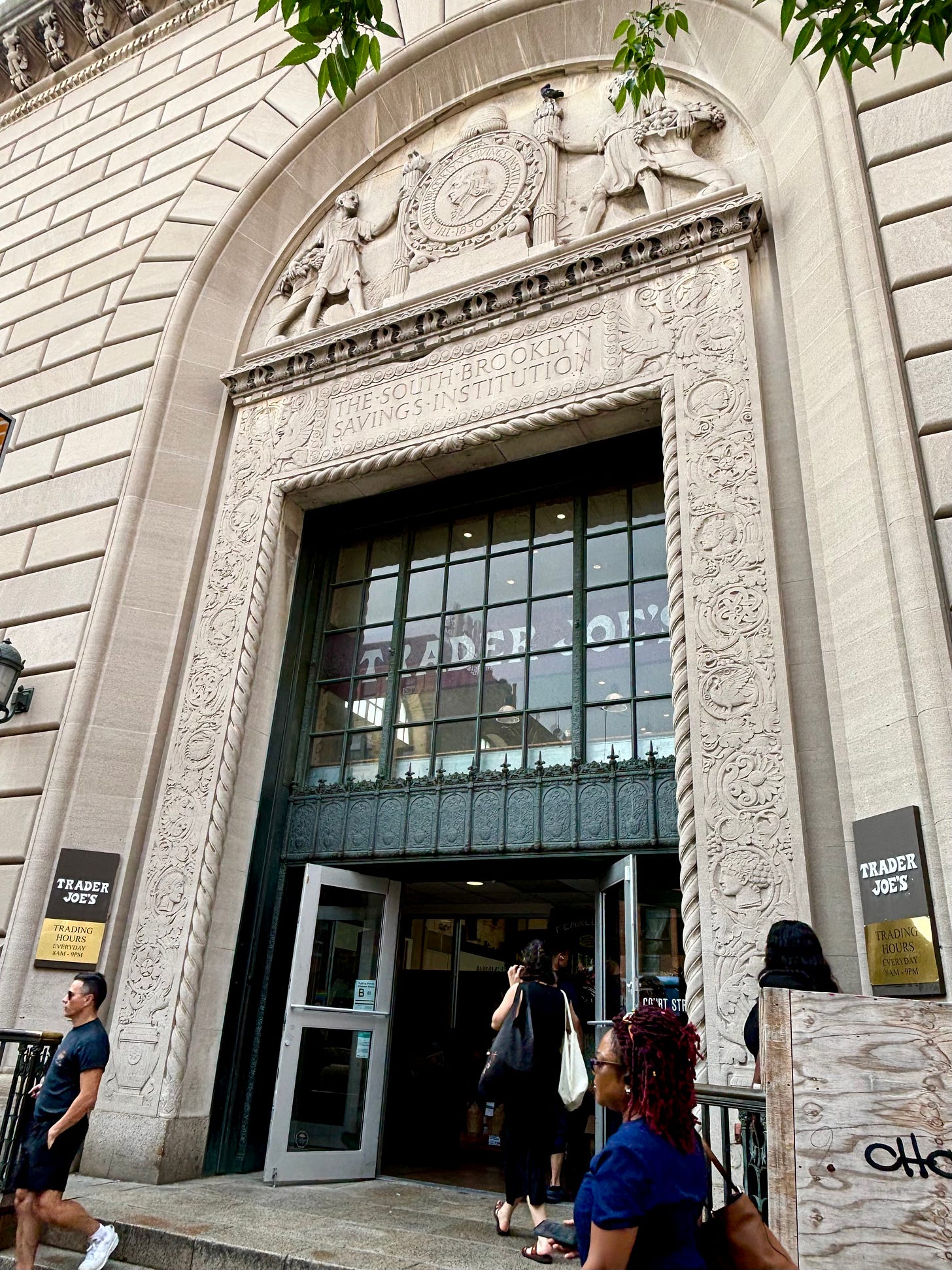An ornately carved arched entrance. Over the door is engraved The South Brooklyn Savings Institution. Inside the glass doors are Trader Joe's flags. An ornately carved arched entrance. Over the door is engraved The South Brooklyn Savings Institution. Inside the glass doors are Trader Joe's flags.