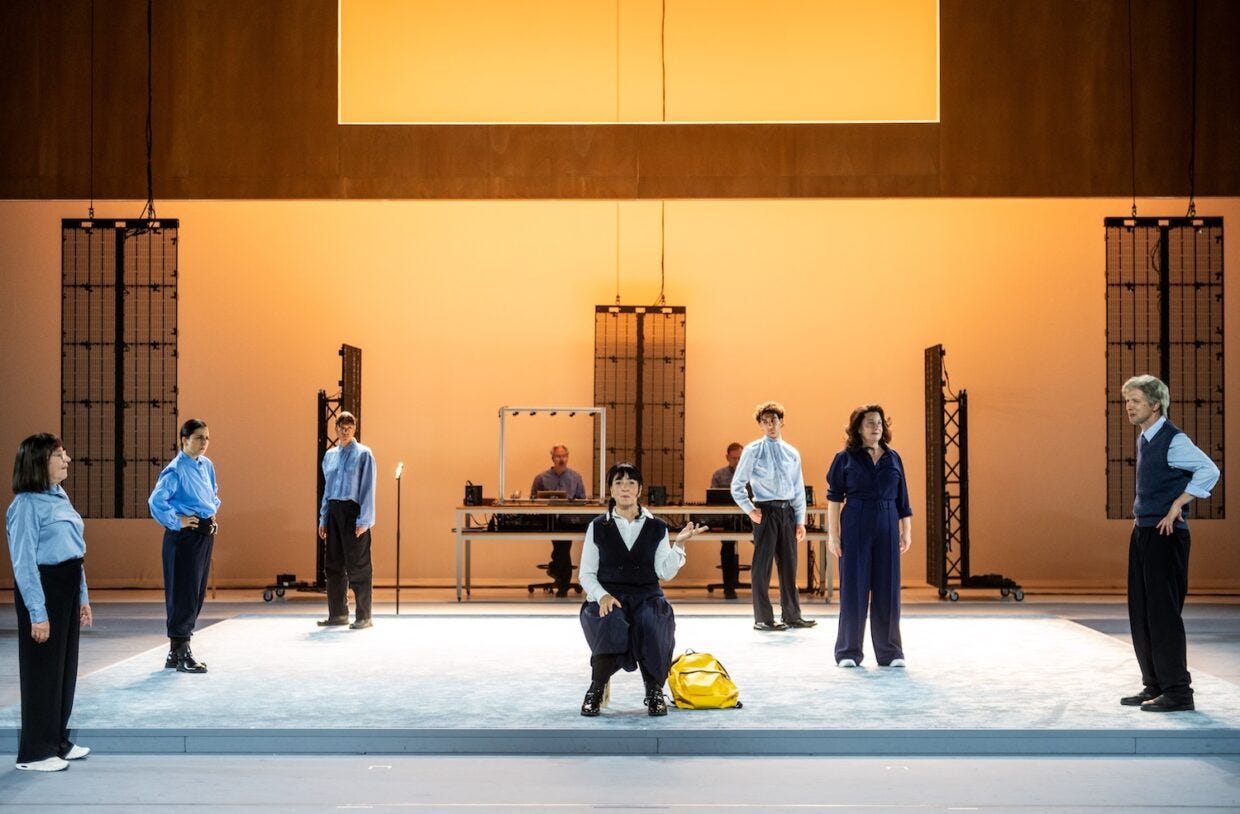 A scene from Spring. The cast, all wearing blue shirts stand around a schoolgirl with a yellow backpack.
