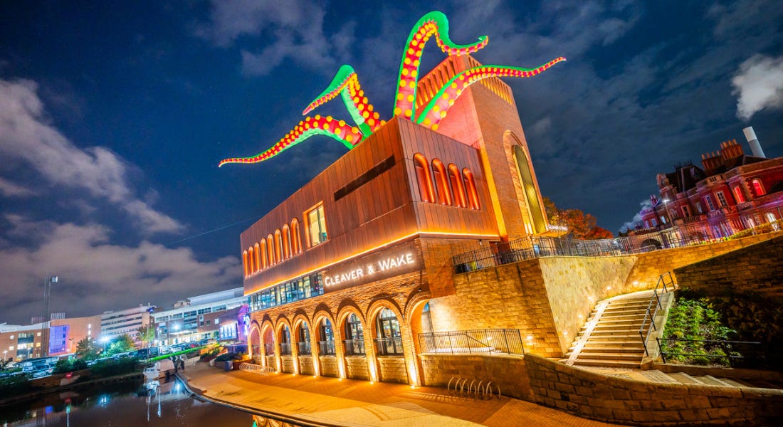 A large wooden and brick building with green tentacles on top of it, with a night sky above it and a river next to it