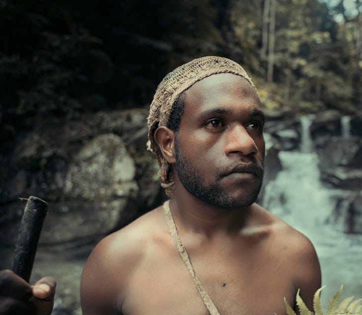 a shirtless man holding a stick in front of a waterfall