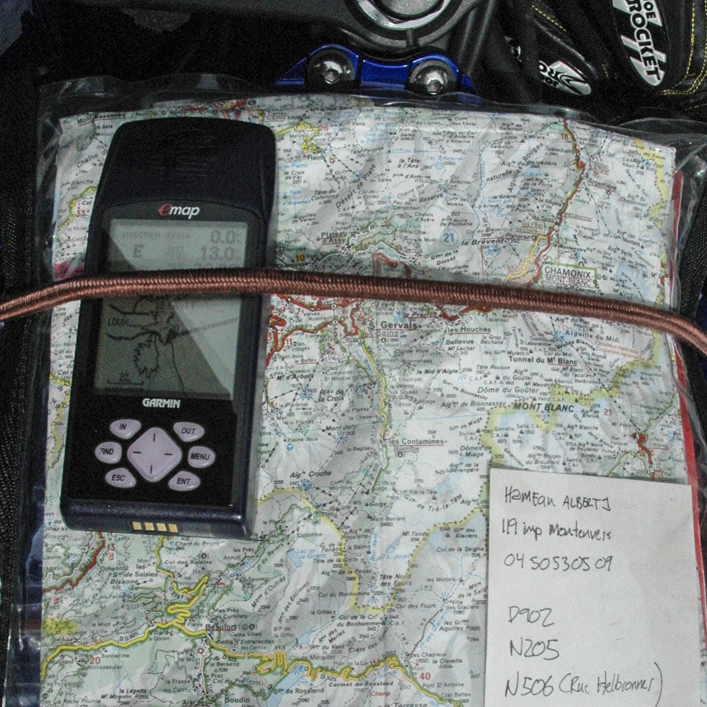 A map pocket mounted to the gas tank of a motorcycle with a bungee cord. The map pocket contains a map, a Garmin emap GPS, and a piece of paper with navigation notes.