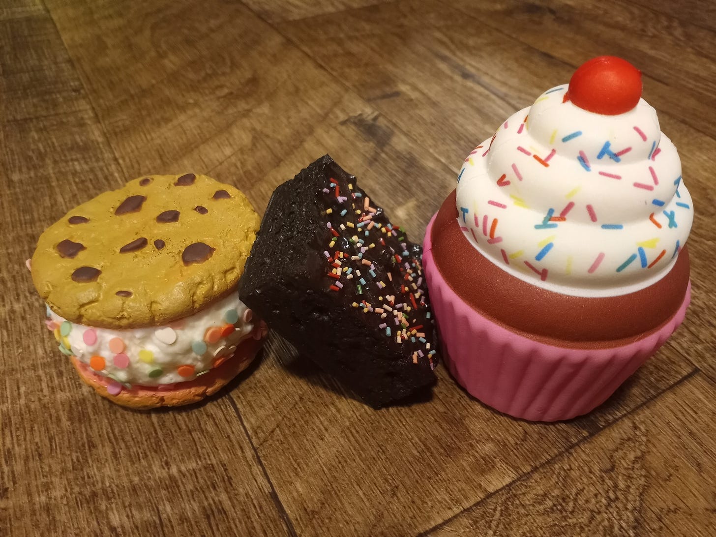 Three realistic dessert-themed squishies, all with rainbow sprinkles. There is a chocolate chip cookie sandwich, a brownie, and a cupcake.