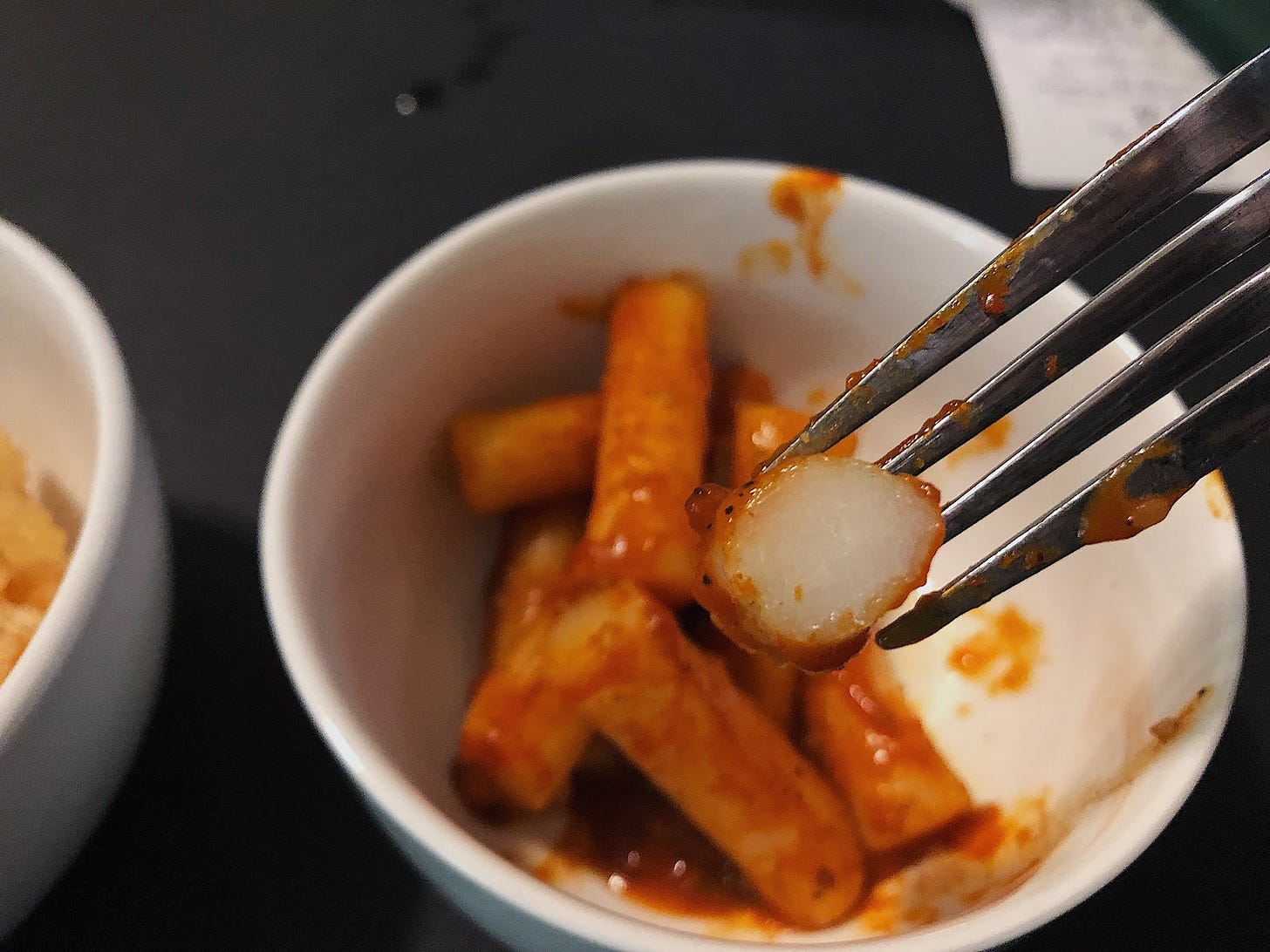 An up close view of a bitten-through piece of Korean rice cake called tteokbokki, covered in a thick sweet and spicy sauce. A small pile of the rice cakes sit in its sauce behind the up close one in a small white bowl.
