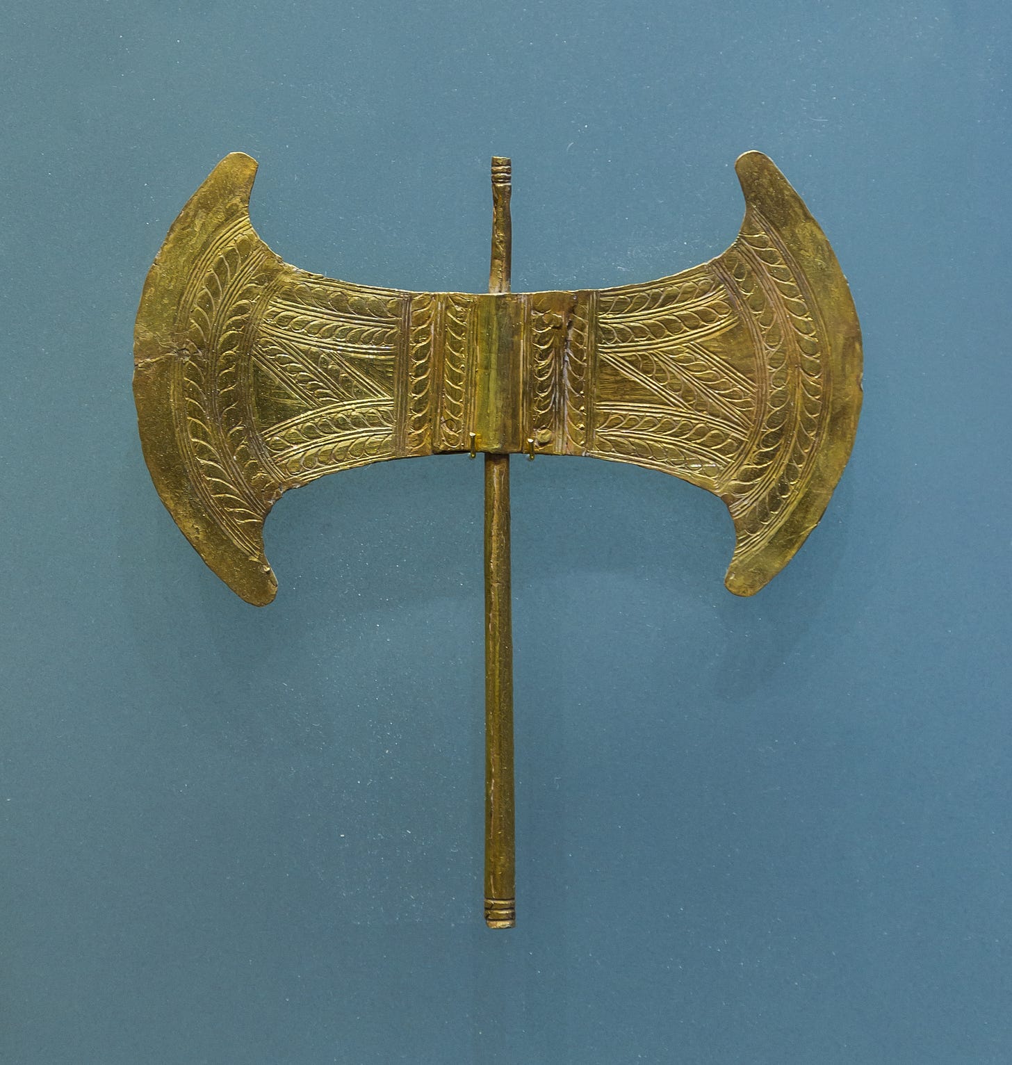 A gold labrys on a light blue background. The labrys is made of thin sheet metal. The blades are incised all over with looping, leaf-like designs. The handle is narrow and shaped like a dowel.