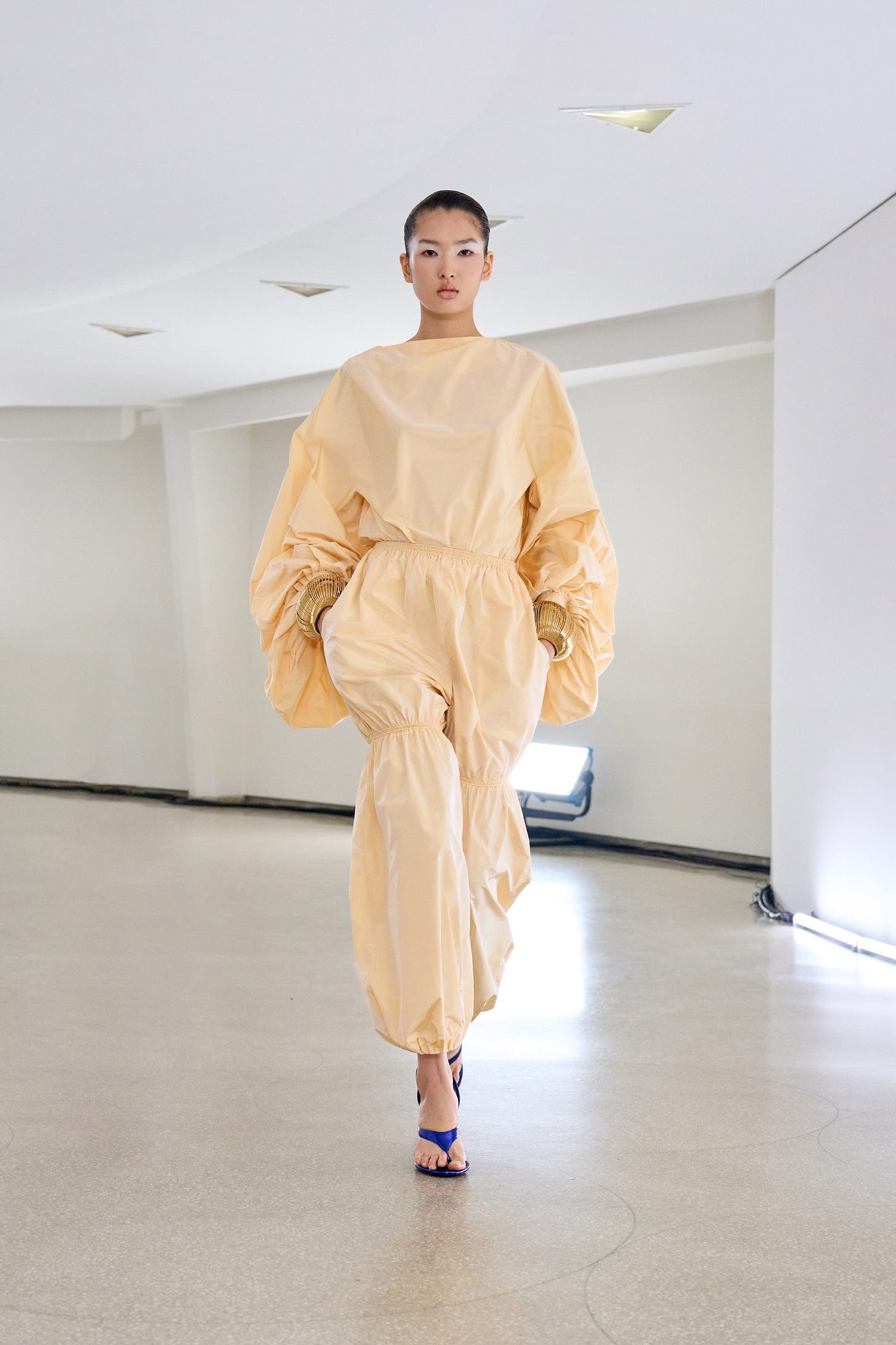 A woman walks down a curved runway wearing a pale yellow top with oversized, gathered sleeves and a matching pant with gathers above the knees.
