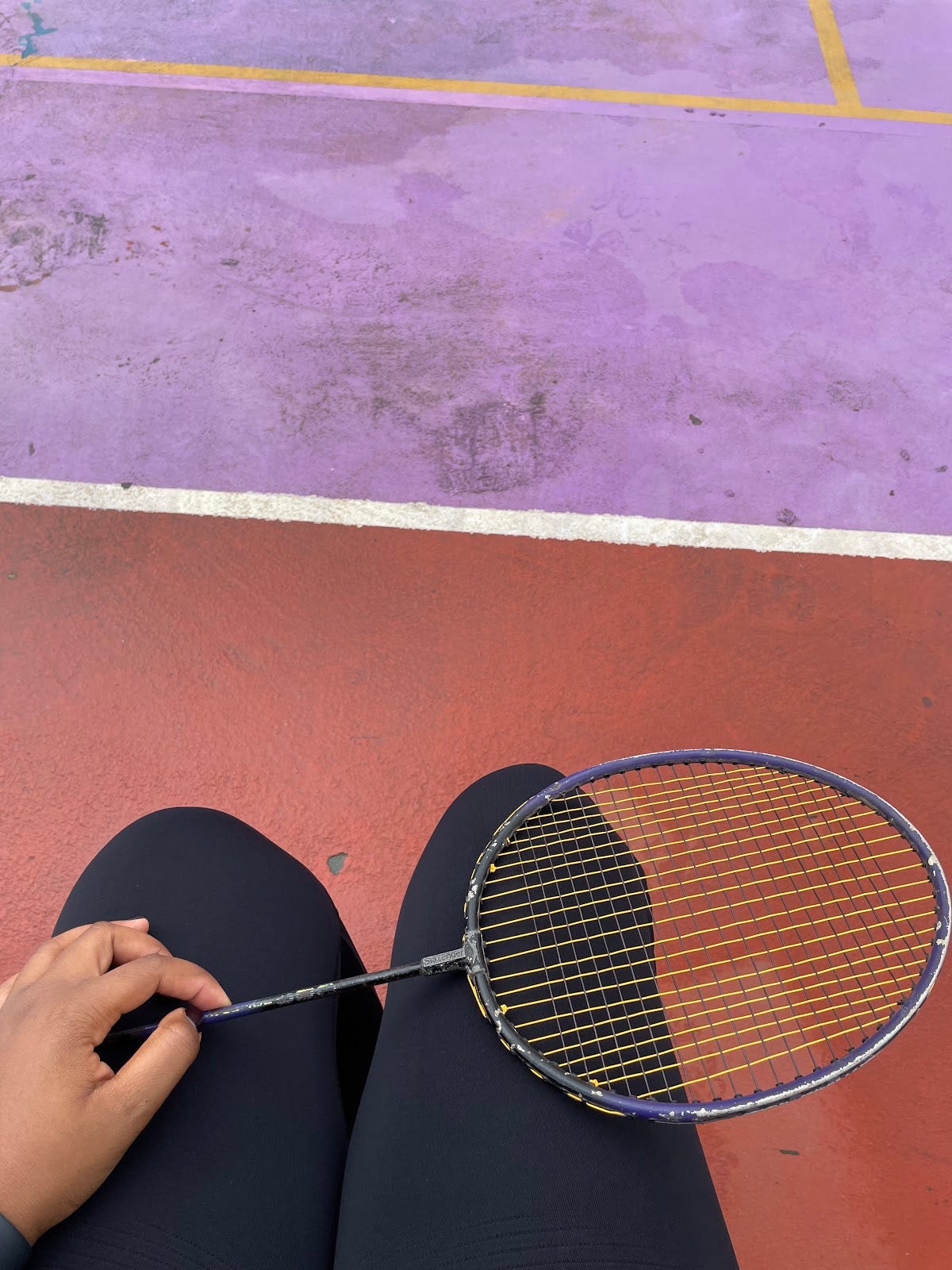 Badminton racket resting on my legs. The floor of the court is purple and brown