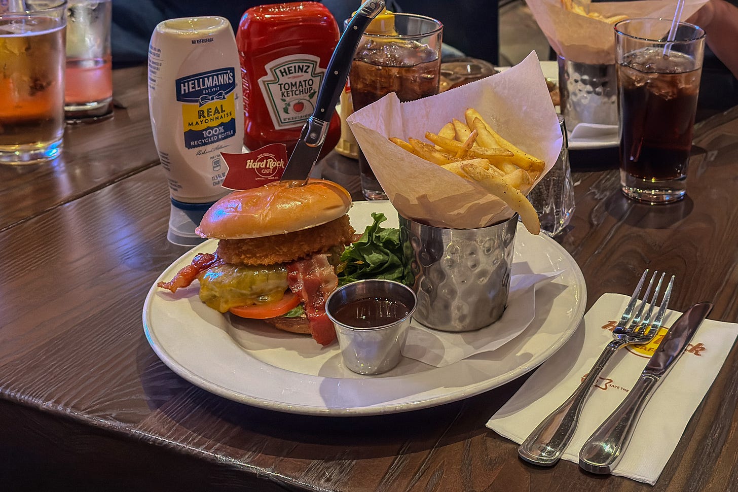 Auf dem dunklen Holztisch in einem Hard Rock Cafe steht ein Teller mit Burger, Pommes frites im Metallbecher und einer Schale Sauce. Im Hintergrund stehen Ketchup, Mayonnaise und mehrere Gläser mit süssen Softdrinks.