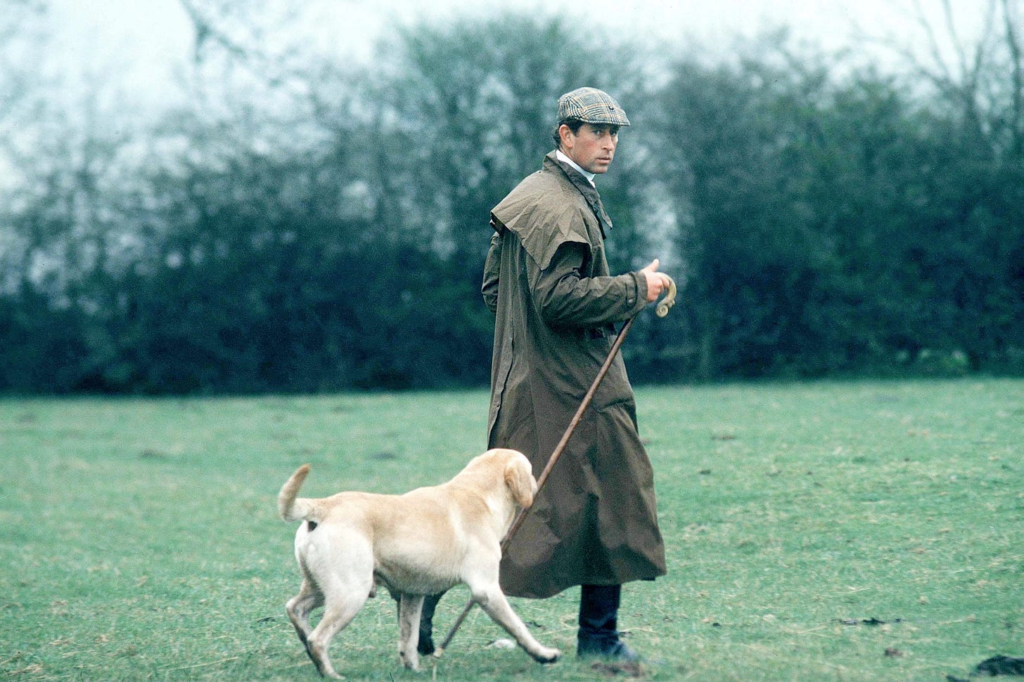 Young King Charles wearing a flat cap and long jacked walking a dog Young King Charles wearing a flat cap and long jacked walking a dog