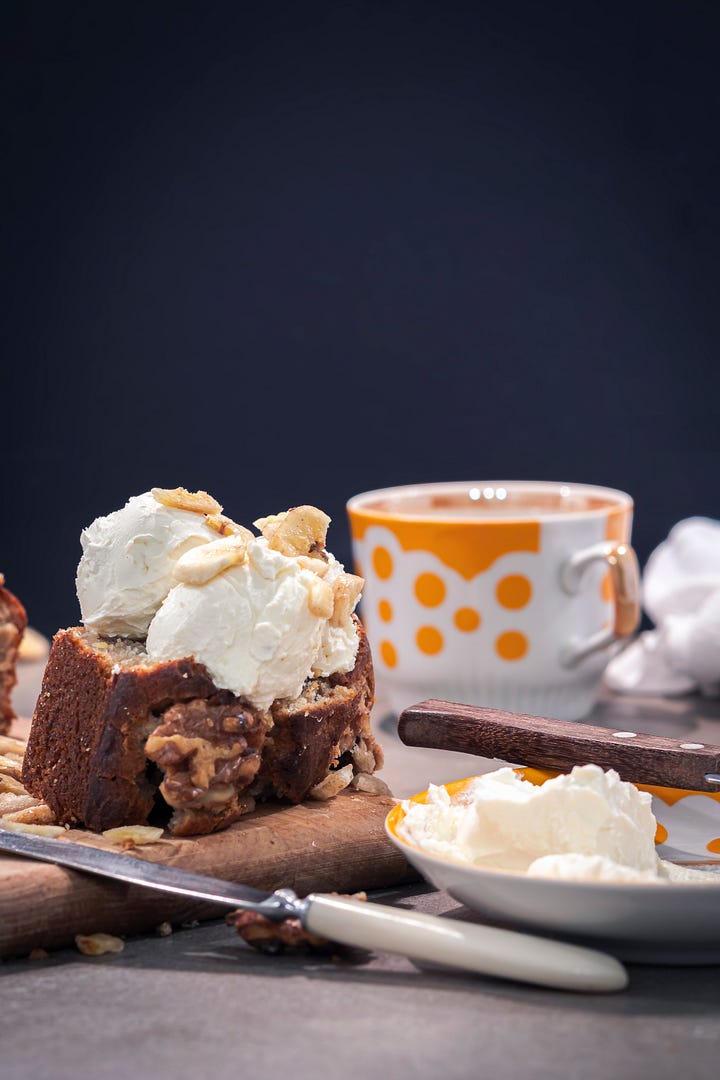 Homemade banana bread topped with pecans, shown whole and sliced, topped with scoops of snow-ball imitating cream cheese. 