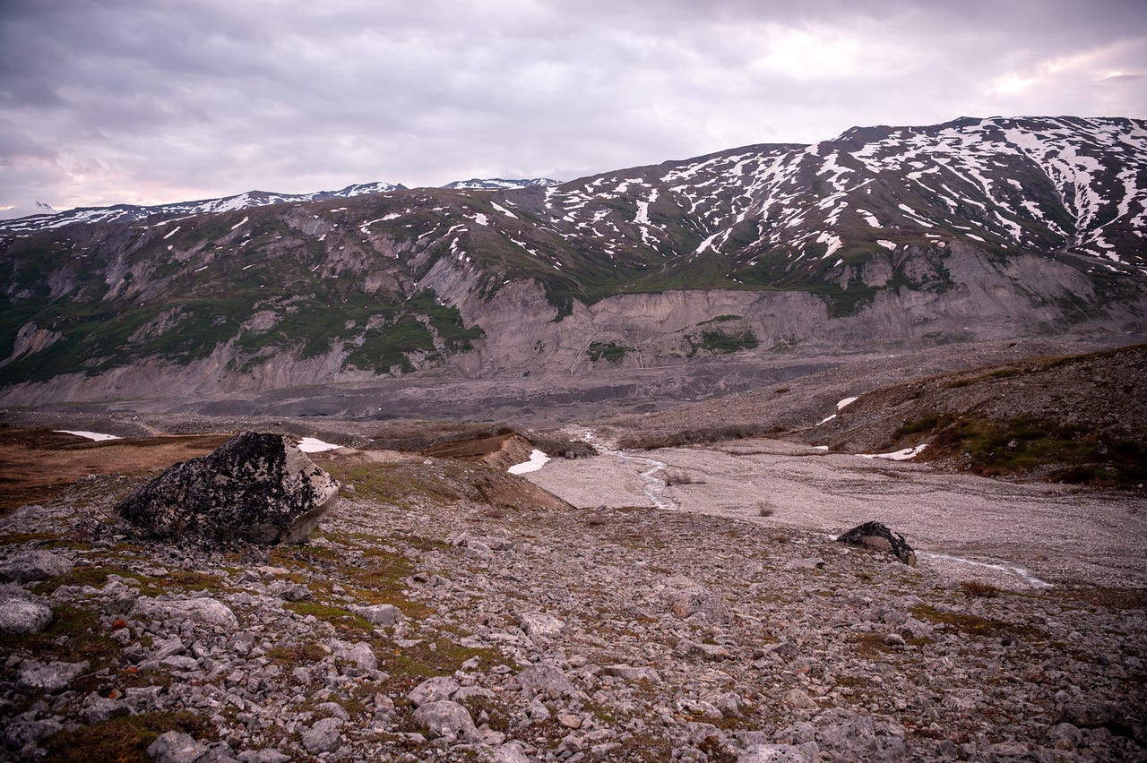Looking across a wide gravel outwash plain threaded by a small braided stream, from a foreground of rocky, sparsely vegetated ground with one large dark boulder. Across the valley, eroded grey bluffs rise to green tundra slopes patched heavily with snow, under a pink-grey dawn sky of soft cloud.