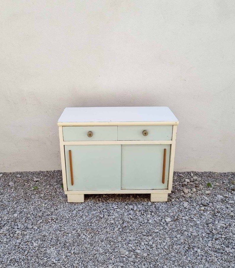 May include: A white cabinet with two drawers and two doors. The cabinet has light blue doors and wooden handles. The cabinet is sitting on a gravel surface.