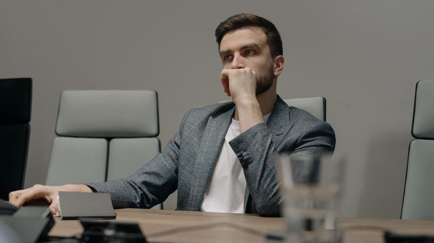 Executive at a boardroom table rests his chin on his hand and looks past the meeting, an empty chair beside him.