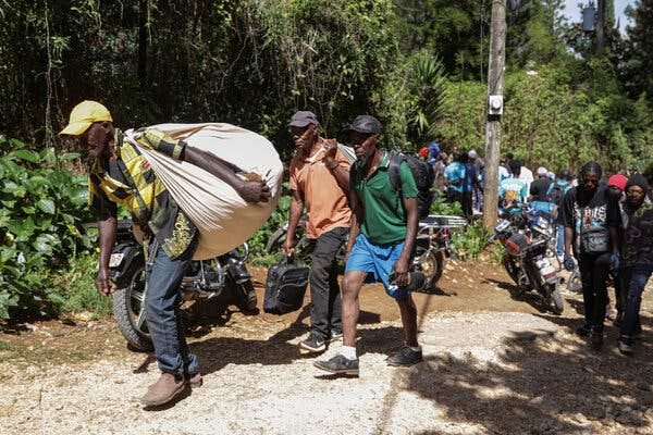 On a sunlit dirt path, a person in a yellow hat and yellow and black shirt carries a large sack over one shoulder. Others walk behind uphill, near motorcycles parked near lush trees.