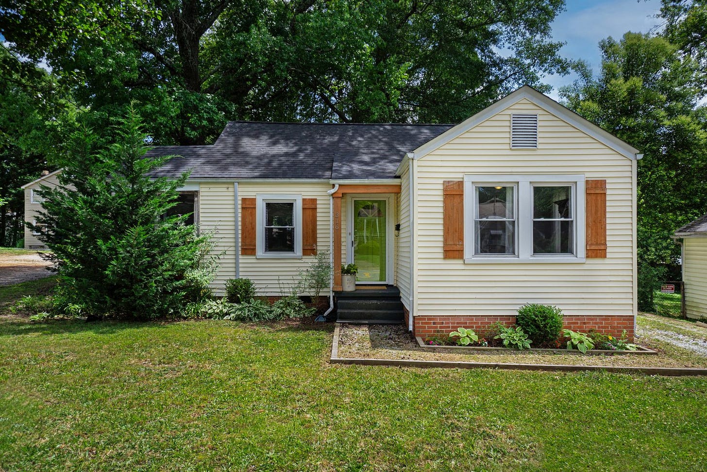 A small cream-colored single-storey house with orange shutters and a brick skirt, with shrubs and a mowed front lawn