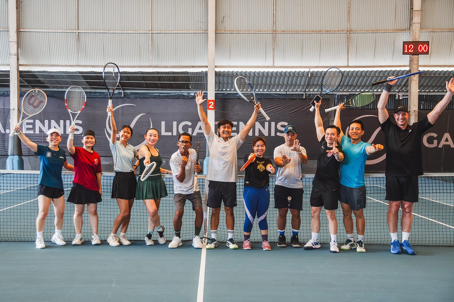 A wide shot of a group of 11 tennis players, men and women of various ethnicities, posing joyfully on an indoor blue tennis court. They are lined up behind a net, many raising their rackets or hands in the air to celebrate. Behind them is a large black banner with "LIGA.TENNIS" printed in white. On the far right, a digital clock shows "12 00". The atmosphere is vibrant and energetic, reflecting the growing community of racquet sports enthusiasts in Indonesia. A wide shot of a group of 11 tennis players, men and women of various ethnicities, posing joyfully on an indoor blue tennis court. They are lined up behind a net, many raising their rackets or hands in the air to celebrate. Behind them is a large black banner with "LIGA.TENNIS" printed in white. On the far right, a digital clock shows "12 00". The atmosphere is vibrant and energetic, reflecting the growing community of racquet sports enthusiasts in Indonesia.