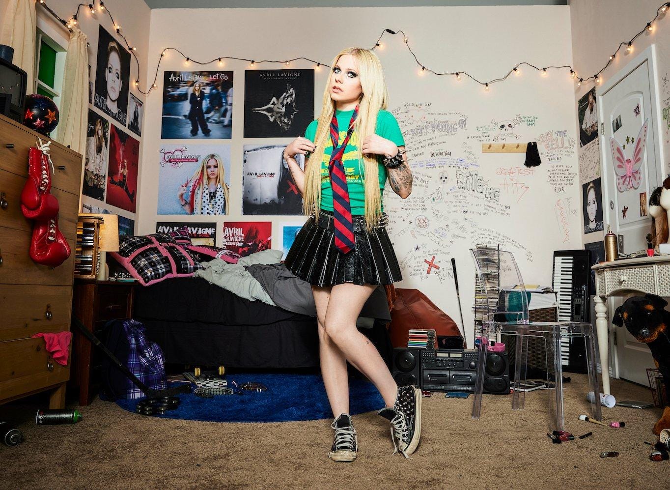 Avril Lavigne in her 30s poses like Avril the teenager: t-shirt with tie, tiny skirt and battered Converse. She stands in a messy bedroom that is covered with posters of every single Avril Lavigne album. There's makeup on the floor, a bedspread spilling onto the ground, a CD tower, a Hi-Fi stereo, and fairy lights. 