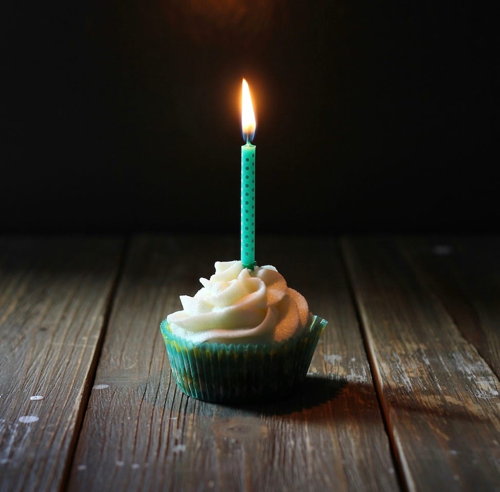 A picture of a lone cupcake with a single candle that appears to light up the area around it in a dark space. The cupcake is in teal and yellow paper; the flavor cannot be determined. The top is covered in what appears to be cream cheese forsting. The single candle is tall and seafoam green with teal polka dots.