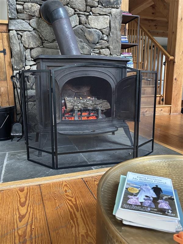 a fire lit in a wood-burning stove in front of a stone chimney. A copy of Elena Ferrante's My Brilliant Friend sits on a metal coffee table in front of the fire.